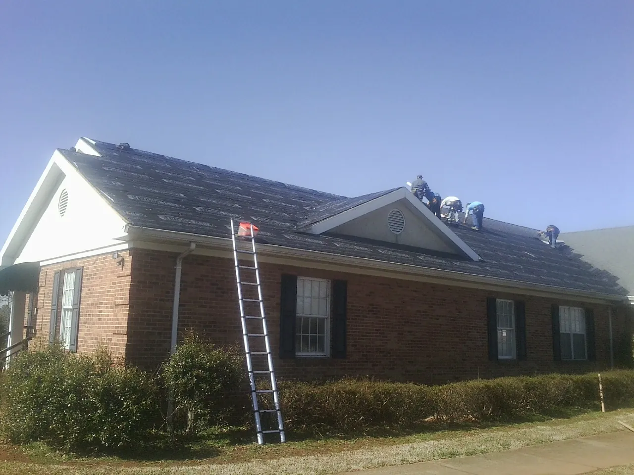 Crew tearing shingles off a brick ranch roof with a ladder set — Emergency Roof Repair work by our crew in Ayden