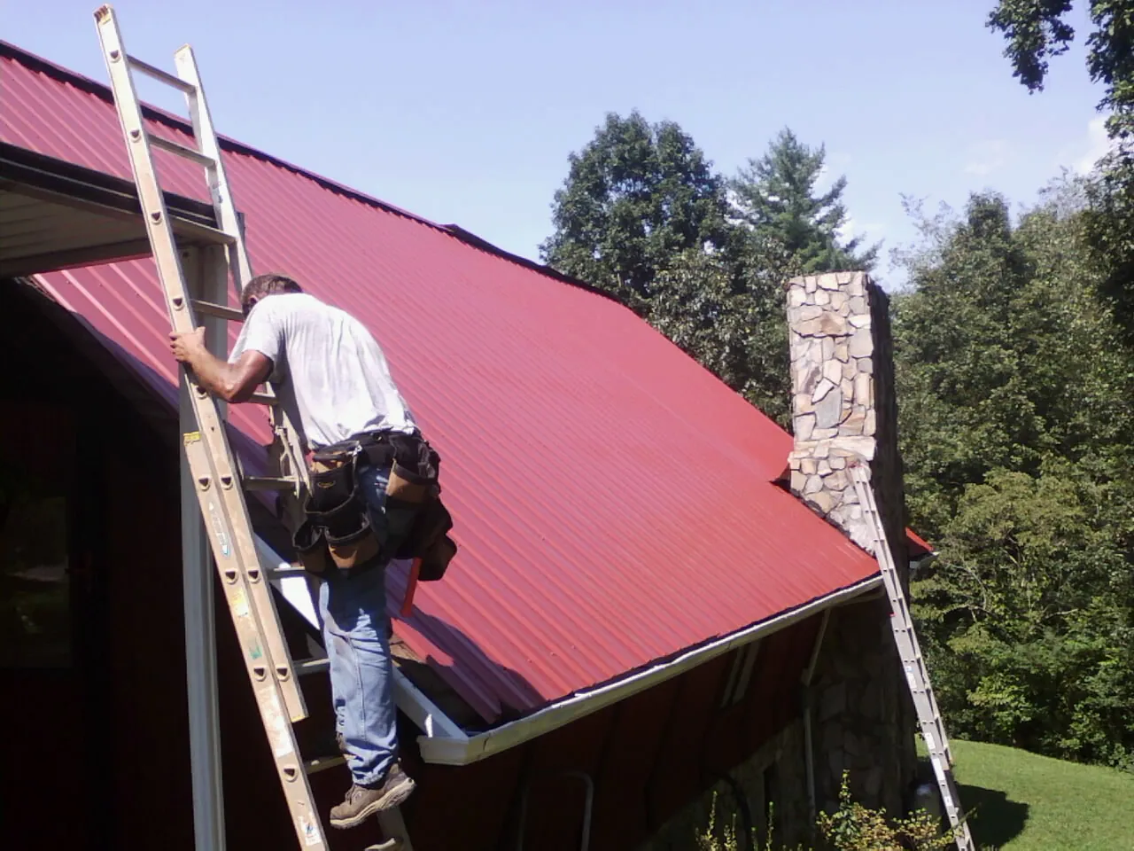 Roofer on a ladder beside a red metal roof and stone chimney — Soffit & Fascia Repair by Roofing Brothers in Ayden